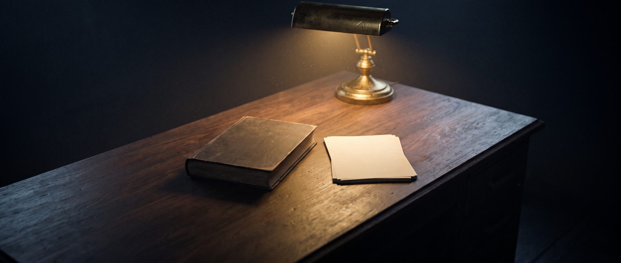 A long dark desk under one lamp, a closed book and loose paper on it